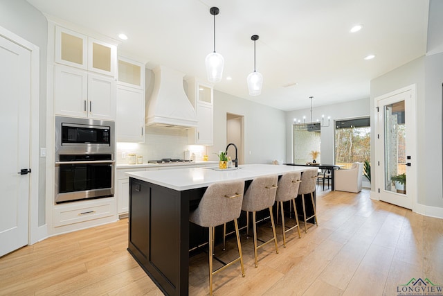 kitchen featuring white cabinets, appliances with stainless steel finishes, custom exhaust hood, an island with sink, and light wood-type flooring