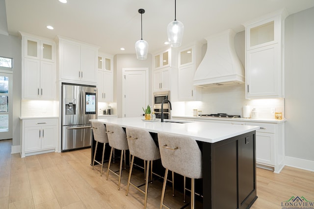 kitchen with custom exhaust hood, appliances with stainless steel finishes, white cabinetry, sink, and a center island with sink