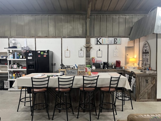 kitchen with black refrigerator with ice dispenser, a sink, concrete flooring, beamed ceiling, and a kitchen breakfast bar