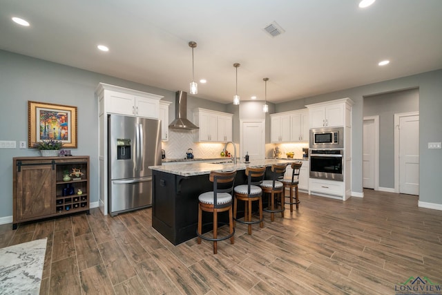 kitchen with pendant lighting, white cabinets, wall chimney exhaust hood, an island with sink, and appliances with stainless steel finishes