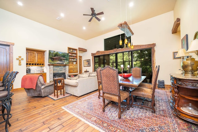 dining area featuring ceiling fan, a stone fireplace, visible vents, a towering ceiling, and light wood finished floors