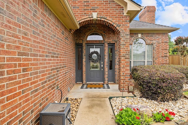 view of exterior entry featuring roof with shingles, a chimney, fence, and brick siding