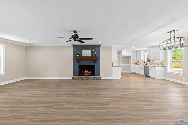 unfurnished living room with crown molding, light wood finished floors, a brick fireplace, baseboards, and ceiling fan with notable chandelier