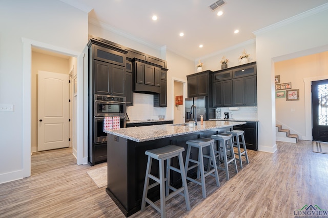 kitchen featuring light wood-type flooring, an island with sink, double oven, tasteful backsplash, and light stone counters