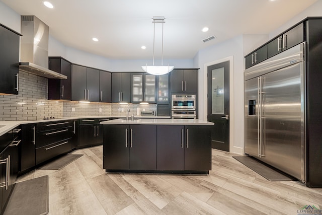 kitchen featuring appliances with stainless steel finishes, sink, hanging light fixtures, a kitchen island with sink, and wall chimney range hood