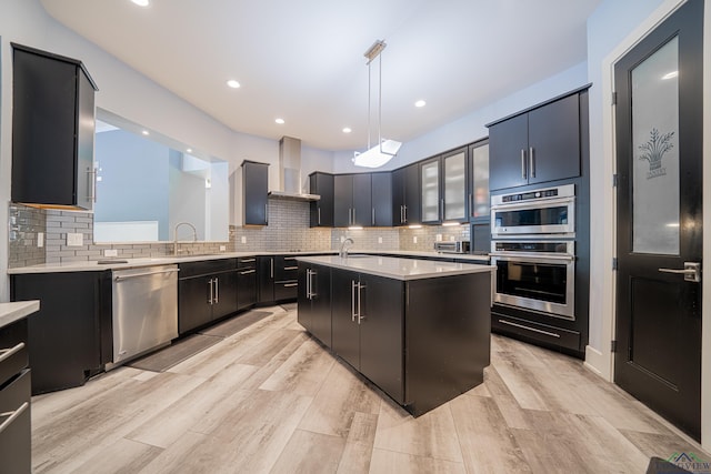 kitchen featuring wall chimney range hood, appliances with stainless steel finishes, a kitchen island with sink, tasteful backsplash, and decorative light fixtures