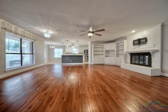 unfurnished living room with crown molding, a brick fireplace, ceiling fan with notable chandelier, and wood-type flooring