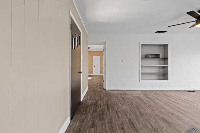 empty room featuring brick wall, a textured ceiling, built in shelves, dark hardwood / wood-style floors, and ceiling fan