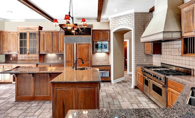 kitchen with built in appliances, decorative backsplash, premium range hood, a kitchen island with sink, and beam ceiling