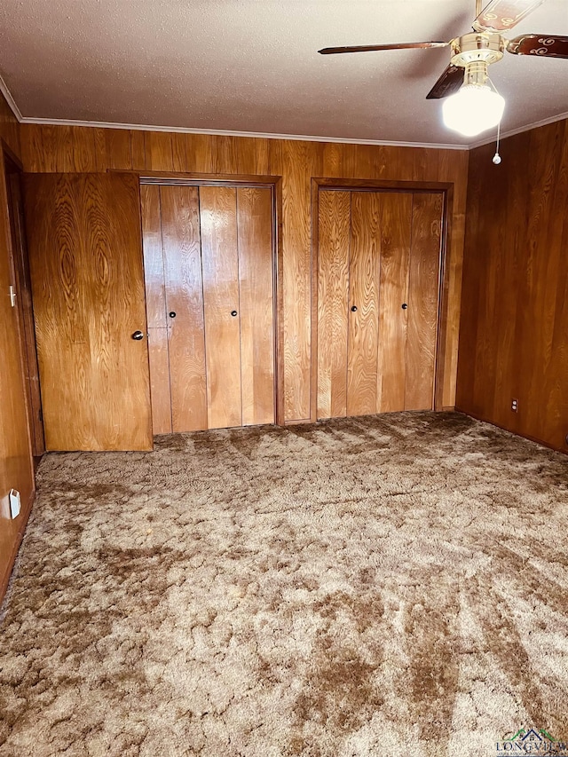 unfurnished bedroom featuring ceiling fan, wooden walls, carpet floors, a textured ceiling, and two closets