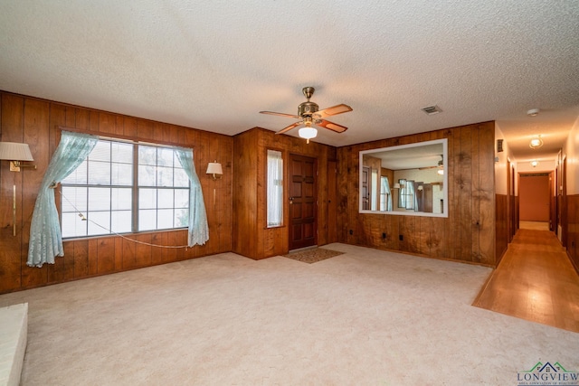 unfurnished living room featuring a textured ceiling, ceiling fan, light colored carpet, and wood walls