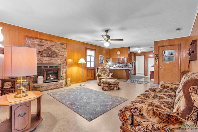 living room featuring wooden walls, visible vents, a ceiling fan, and a brick fireplace