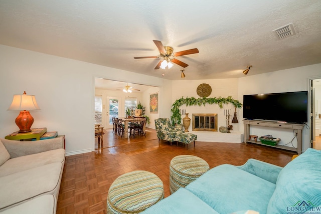 living room with a textured ceiling, a brick fireplace, ceiling fan, and dark parquet floors