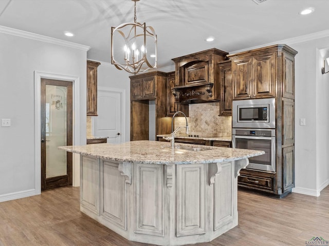 kitchen with light stone countertops, appliances with stainless steel finishes, ornamental molding, a kitchen island with sink, and a notable chandelier