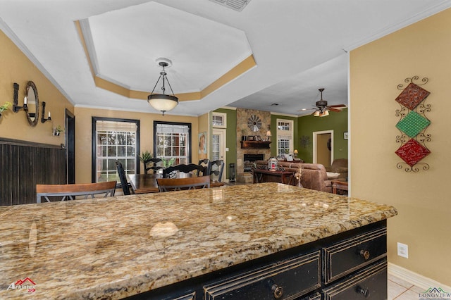 kitchen with light tile patterned flooring, a stone fireplace, pendant lighting, a raised ceiling, and crown molding