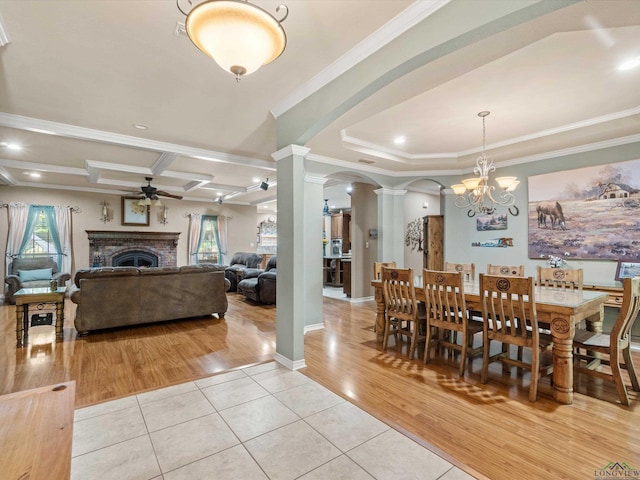 tiled dining space featuring ceiling fan with notable chandelier, decorative columns, ornamental molding, and a brick fireplace