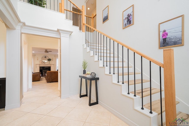 staircase featuring baseboards, a high ceiling, a fireplace, ceiling fan, and tile patterned floors