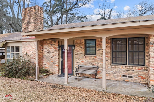 view of exterior entry with crawl space, brick siding, a chimney, and a porch