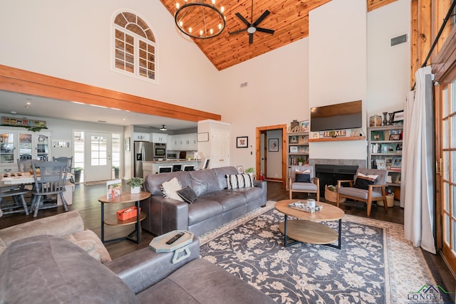 living room featuring wooden ceiling, high vaulted ceiling, wood-type flooring, a tiled fireplace, and ceiling fan with notable chandelier