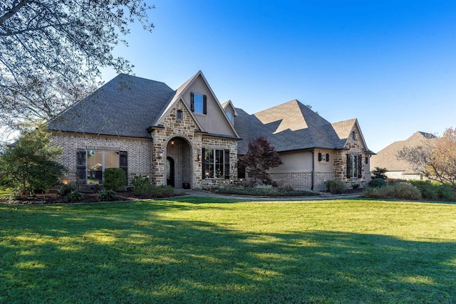 french country style house featuring a shingled roof, a front yard, stone siding, and stucco siding