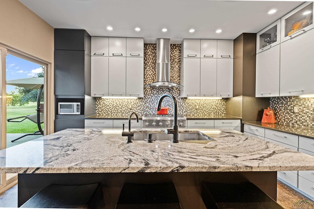 kitchen featuring wall chimney exhaust hood, light stone counters, a breakfast bar area, and a large island with sink