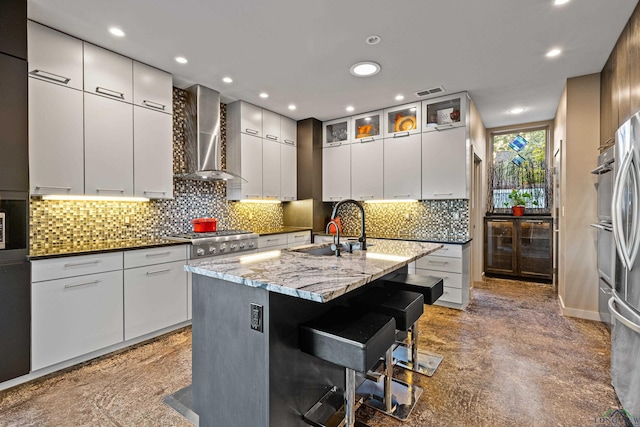 kitchen featuring white cabinets, a kitchen breakfast bar, wall chimney range hood, sink, and an island with sink