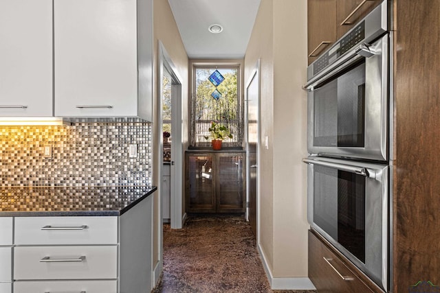 kitchen with white cabinets, dark stone countertops, stainless steel double oven, and tasteful backsplash