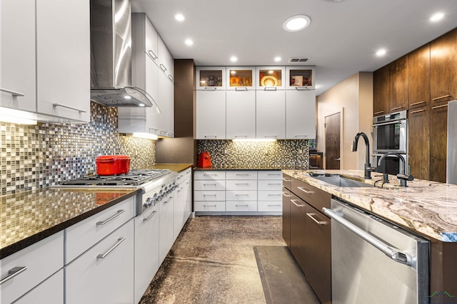 kitchen featuring wall chimney range hood, sink, light stone countertops, appliances with stainless steel finishes, and white cabinetry