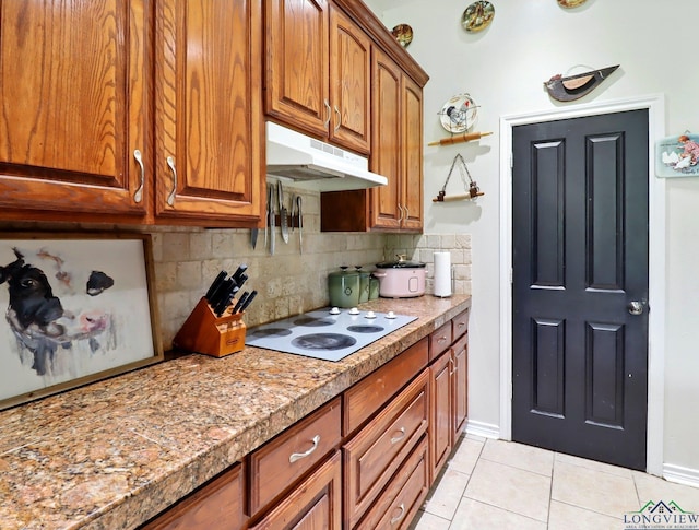 kitchen with white electric stovetop, brown cabinets, light tile patterned flooring, under cabinet range hood, and backsplash