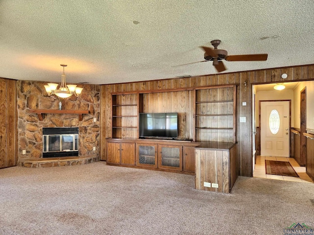 unfurnished living room with light carpet, a textured ceiling, wooden walls, a fireplace, and ceiling fan with notable chandelier