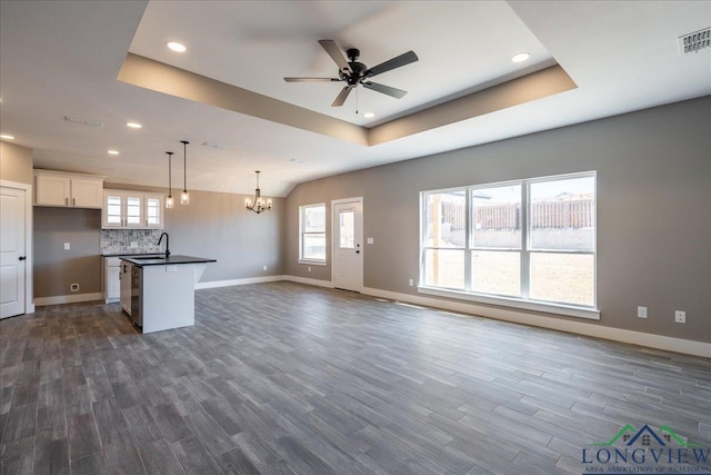 unfurnished living room with dark wood-type flooring, ceiling fan with notable chandelier, a tray ceiling, and sink