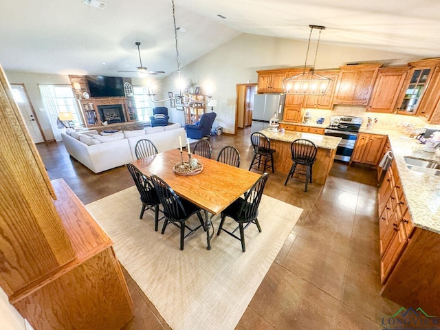 dining room with ceiling fan with notable chandelier, a stone fireplace, sink, and vaulted ceiling