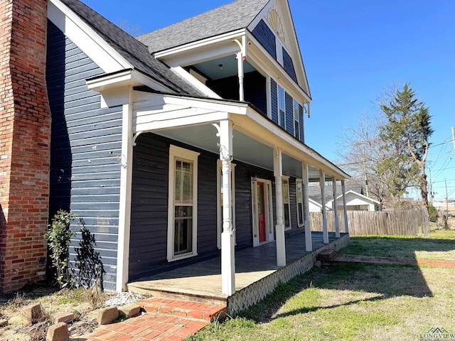 view of side of home featuring a lawn, covered porch, roof with shingles, and fence