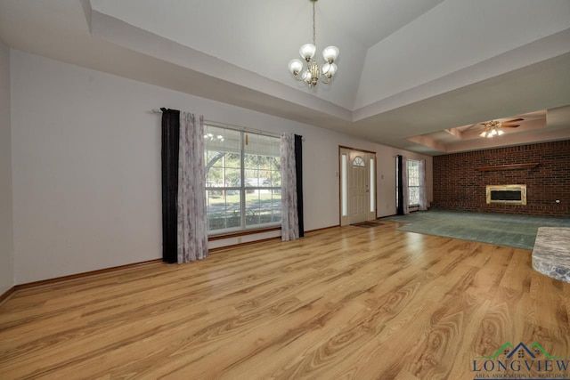 unfurnished living room featuring a brick fireplace, ceiling fan with notable chandelier, light wood-type flooring, and a tray ceiling