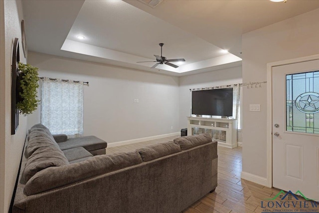 living room featuring baseboards, light wood-style flooring, a tray ceiling, and ceiling fan