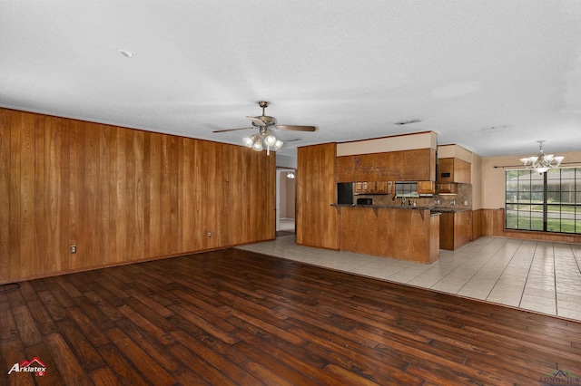 unfurnished living room with a textured ceiling, light wood-style flooring, wooden walls, and ceiling fan with notable chandelier