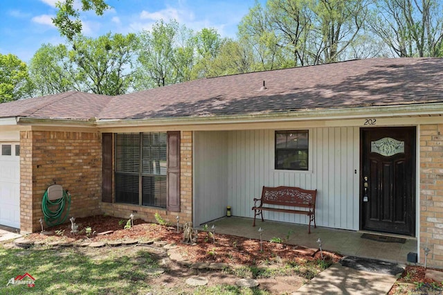 doorway to property with a shingled roof, a porch, a garage, and brick siding