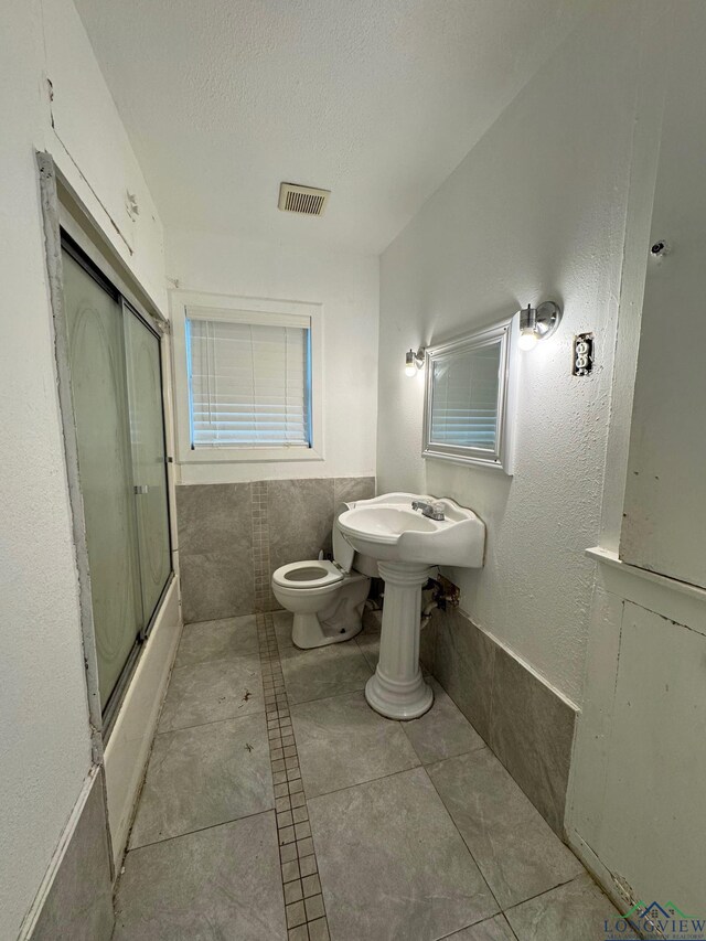 308 E 14th St, Mount Pleasant, TX 75455 - bathroom with tile walls, a textured ceiling, visible vents, toilet, and shower / bath combination with glass door