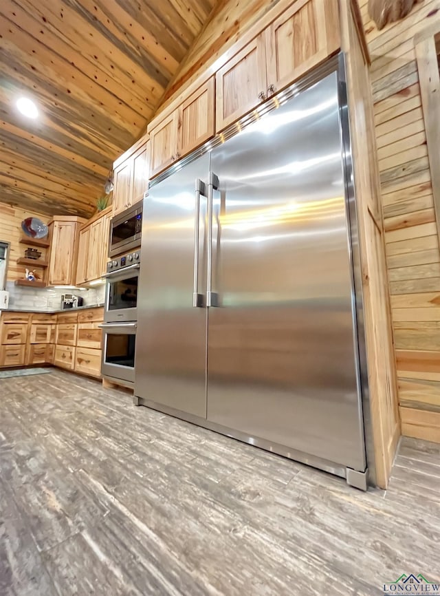 kitchen featuring light brown cabinetry, built in appliances, vaulted ceiling, wooden ceiling, and wooden walls