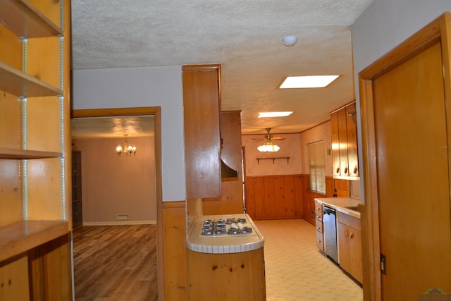 kitchen featuring ceiling fan with notable chandelier, wood walls, hanging light fixtures, stainless steel dishwasher, and white gas cooktop