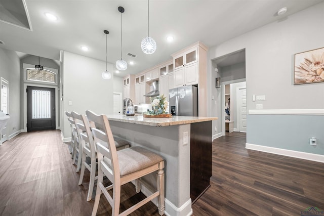 kitchen with wall chimney range hood, stainless steel refrigerator with ice dispenser, decorative light fixtures, light stone counters, and glass insert cabinets