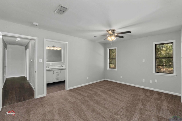 unfurnished bedroom with ensuite bath, ceiling fan, dark carpet, and multiple windows