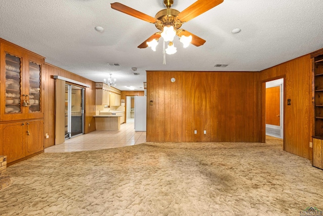 unfurnished living room with light colored carpet, a textured ceiling, ceiling fan with notable chandelier, visible vents, and wooden walls