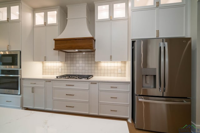 kitchen with white cabinets, decorative backsplash, light stone countertops, appliances with stainless steel finishes, and premium range hood