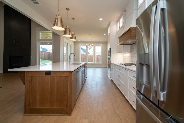 kitchen featuring a large island with sink, a sink, brown cabinetry, appliances with stainless steel finishes, and premium range hood