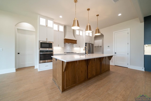 kitchen with stainless steel appliances, a large island with sink, backsplash, custom range hood, and light countertops