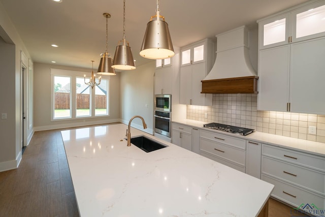 kitchen with an island with sink, backsplash, a sink, appliances with stainless steel finishes, and custom range hood