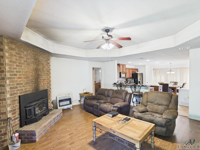 living room with a wood stove, ceiling fan, wood finished floors, a raised ceiling, and heating unit
