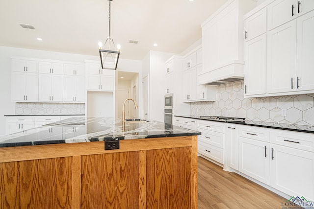 kitchen with decorative light fixtures, stainless steel appliances, dark stone countertops, premium range hood, and a center island with sink