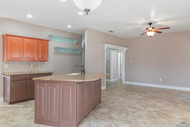 kitchen with sink, decorative backsplash, light stone counters, ceiling fan, and a center island with sink